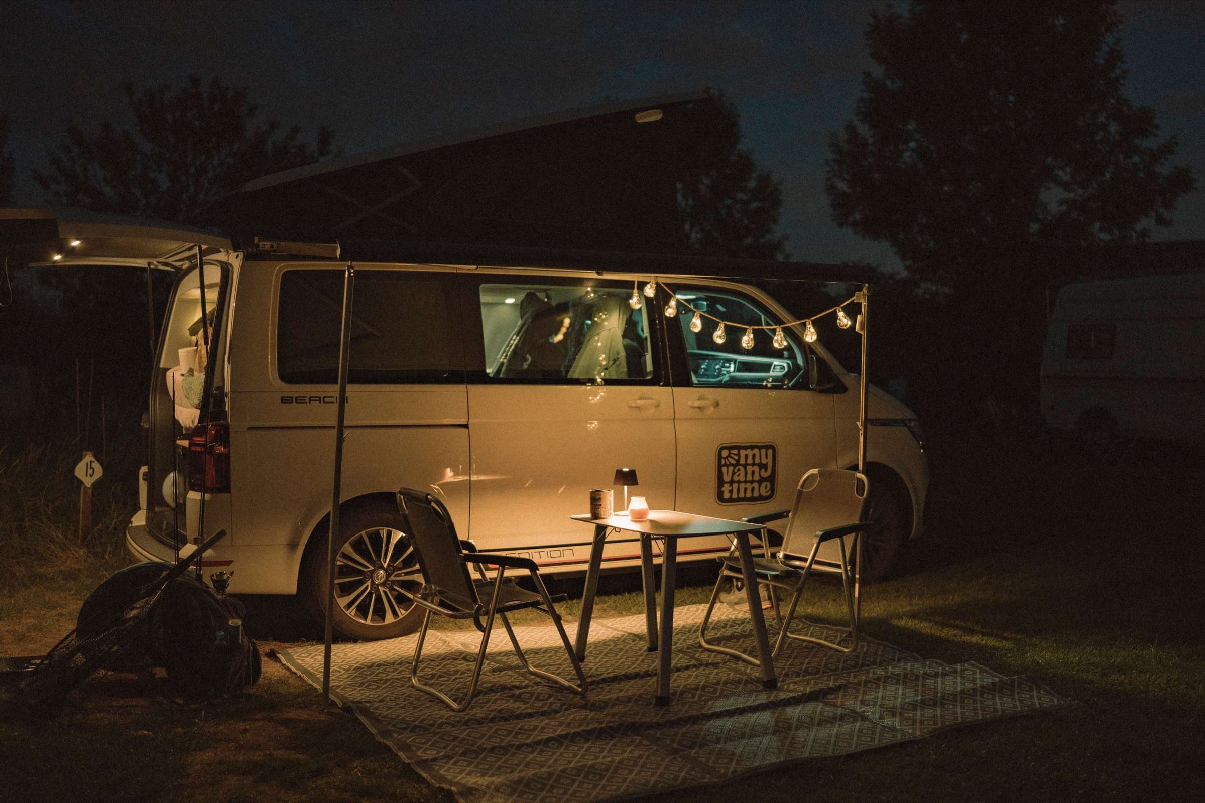 VW T6.1 California Beach am Abend auf dem Stellplatz mit Lichterkette, Tisch und Stühlen unter der Markise